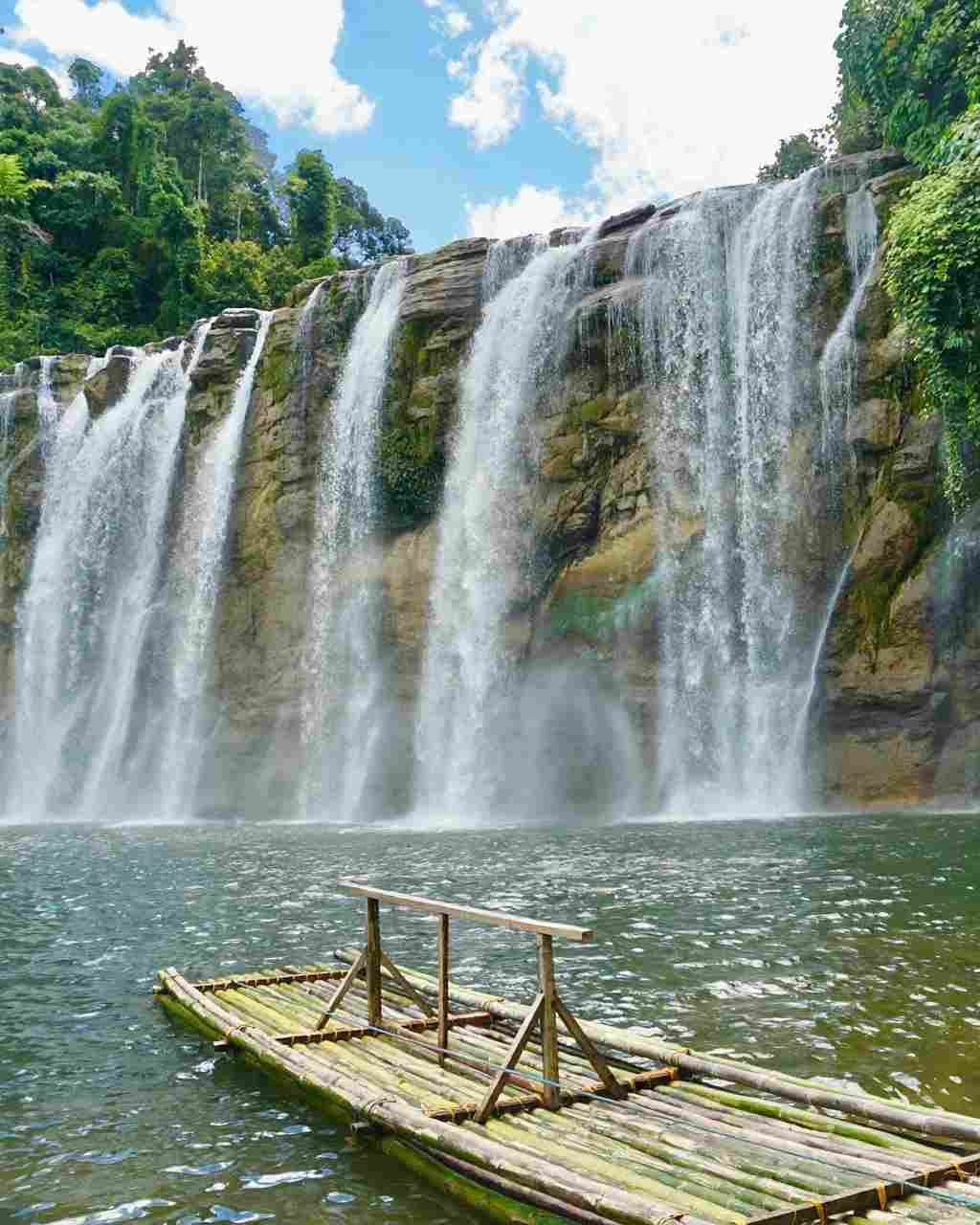 Wide curtain of Tinuy-an Falls in Bislig spilling over a rock cliff into a green pool, with an empty bamboo raft in the foreground
