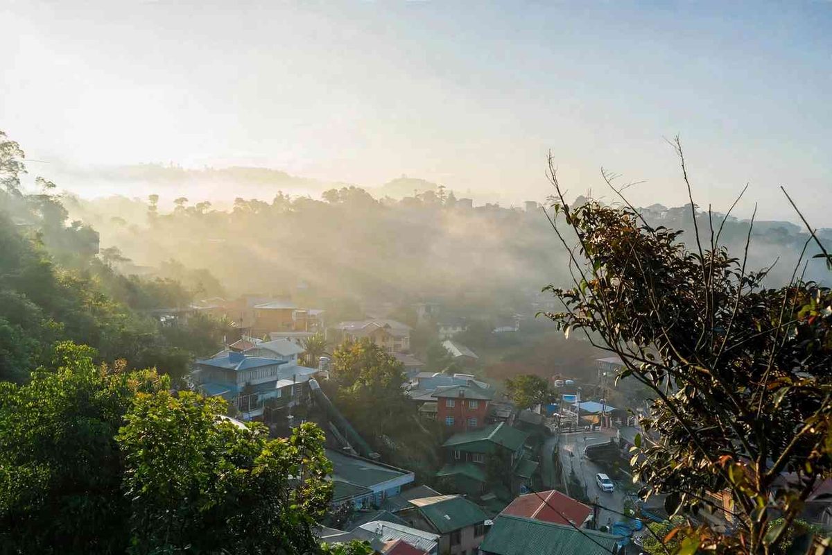 Morning fog drifting over pine-covered hills and houses in Baguio City during the cold season, with sunlight breaking through the mist