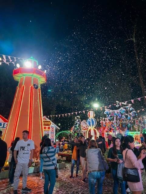 Visitors under falling artificial snow at the Baguio Country Club Christmas Village at night, beside a lit-up red and orange lighthouse and candy-themed decorations