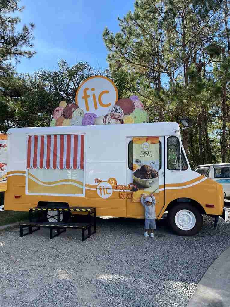 Orange and white ice cream truck parked on gravel under tall pine trees in Baguio, with a child standing by the door