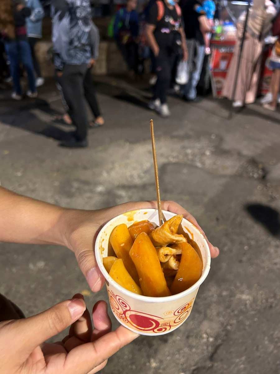 Paper cup of spicy rice cakes and fish cake in red sauce with a wooden skewer at the Baguio Night Market