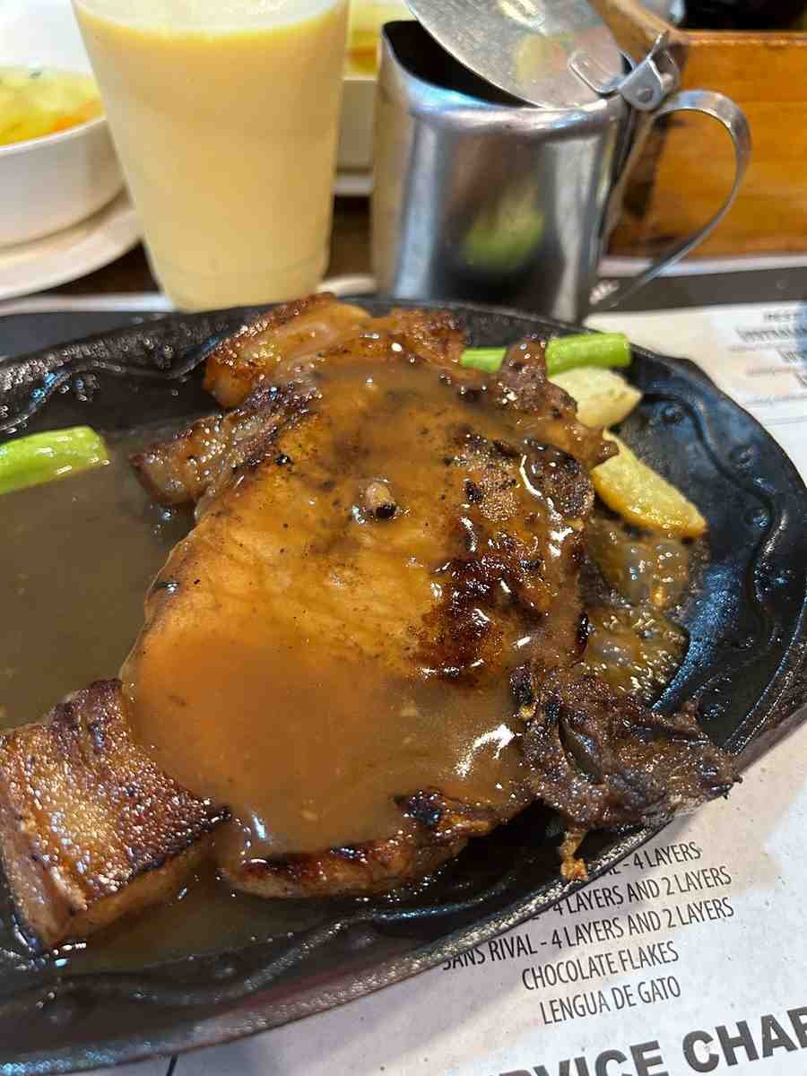 Sizzling pork chop topped with brown gravy on a black iron plate at Sizzling Plate in Baguio, with a glass of fruit shake behind