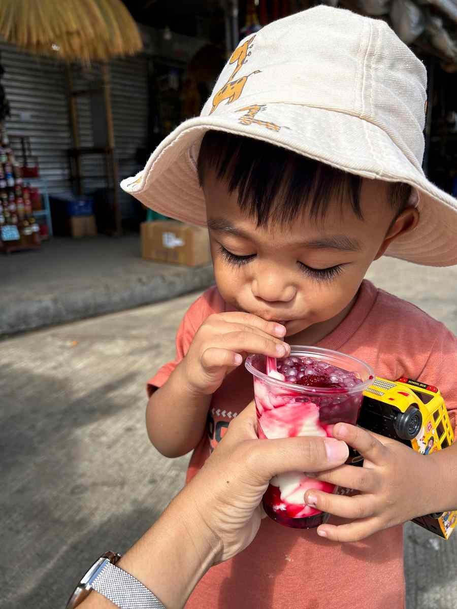 Small boy in a bucket hat sipping strawberry taho through a straw on a Baguio street