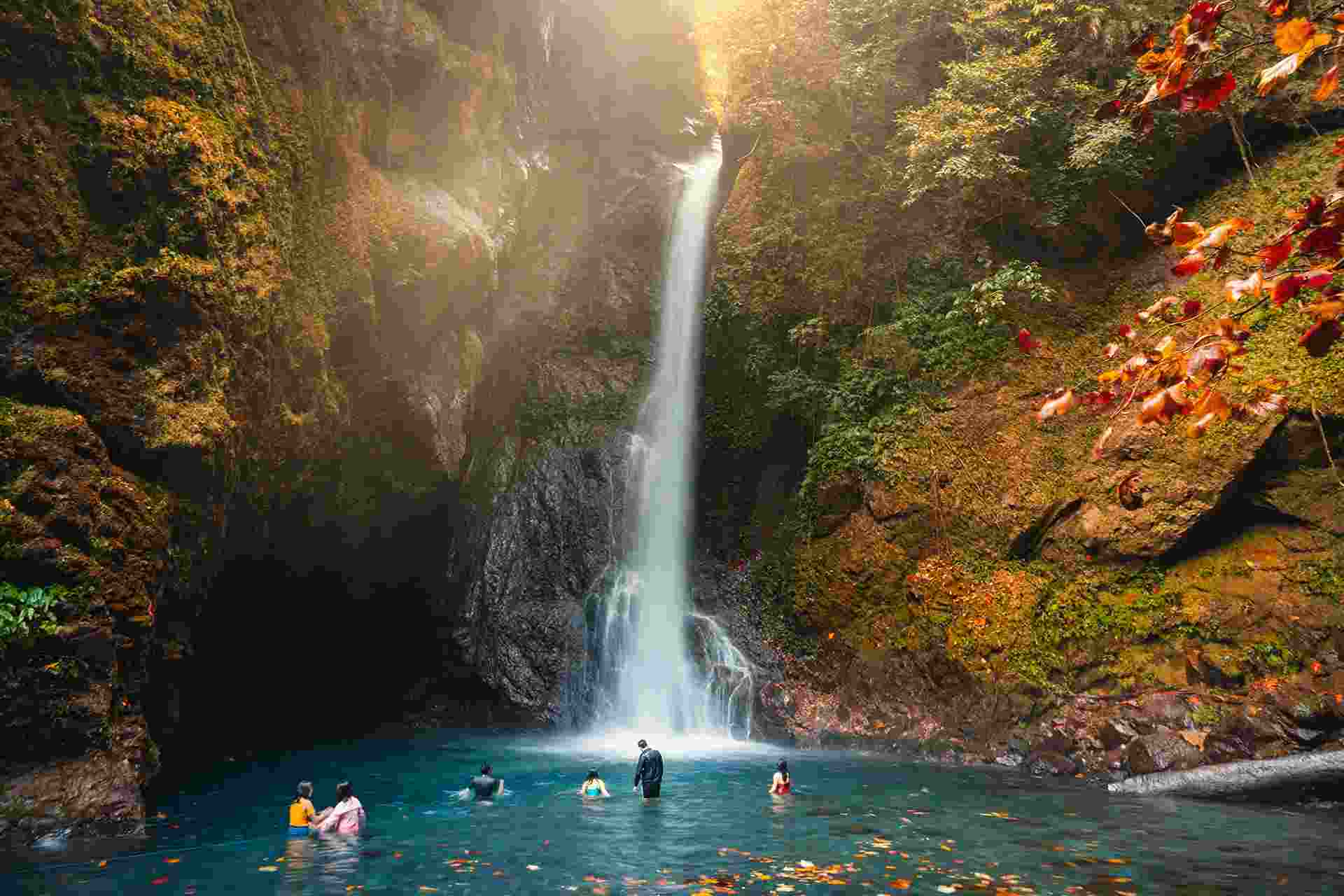 A tall waterfall pouring into a turquoise pool, with sunlight breaking through the mist and swimmers standing in the water at its base