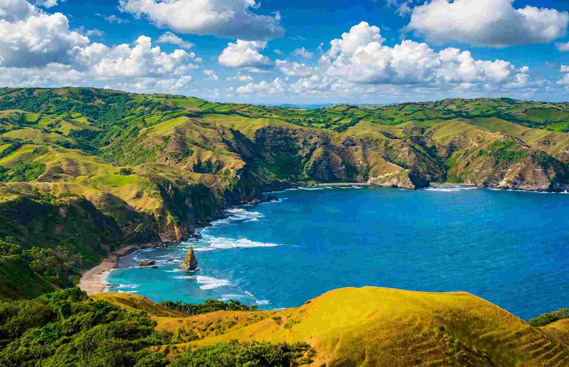 Green rolling hills and a deep blue cove in Batanes, with a tall sea stack standing offshore and white surf breaking on the rocks