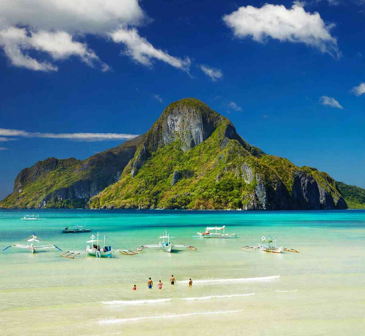 Outrigger bangka boats anchored in shallow turquoise water off a white sand beach in El Nido, Palawan, with the forested limestone peak of Cadlao Island under a deep blue sky