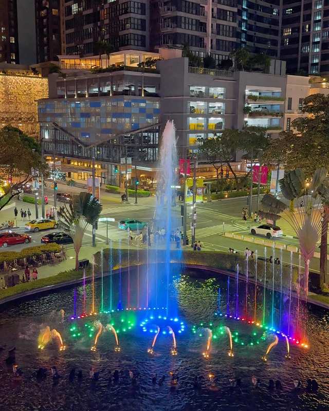 Colourful lit fountain at night in Bonifacio Global City, Taguig, with high-rise buildings, a busy intersection and yellow taxis behind it