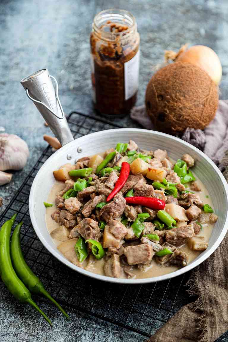Bicol Express of pork, sliced green chilies and red chilies in coconut milk in a white pan, with a jar of shrimp paste, a coconut and garlic beside it