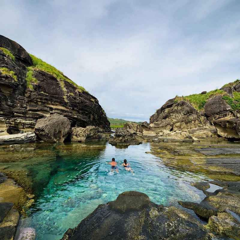 Two swimmers in a clear green-blue tide pool between layered dark rock walls topped with grass at the Biri Island rock formations, Northern Samar