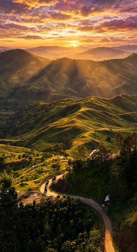 Winding mountain road through green grassy highlands at sunrise, with motorcycles on a bend and layered hills under a golden sky