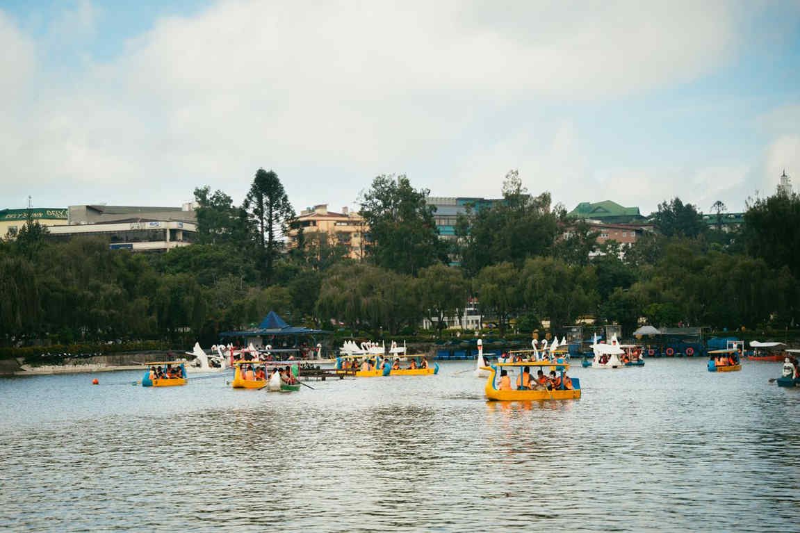 Yellow and white rental boats with passengers in life vests on Burnham Lake in Baguio, with trees and city buildings along the shore