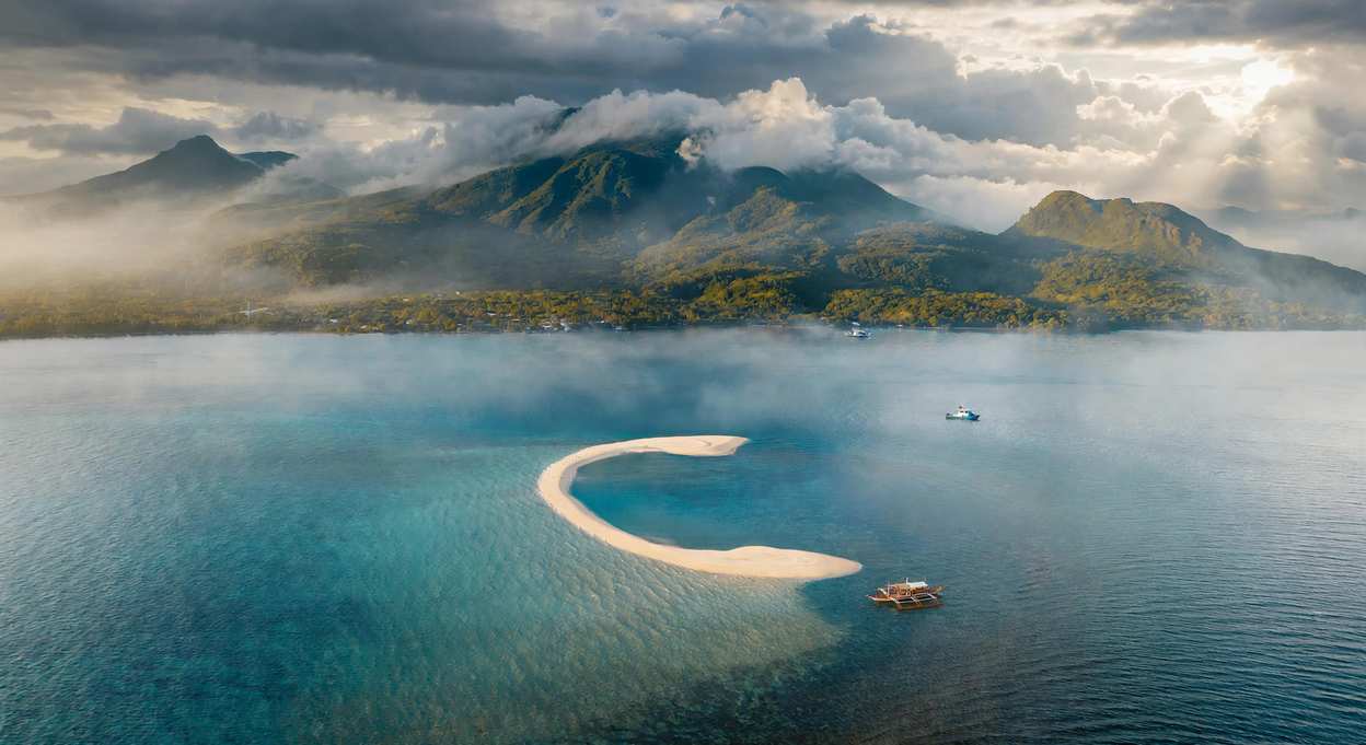 Aerial view of the crescent-shaped White Island sandbar off Camiguin, with outrigger boats on clear water and cloud-covered volcanic peaks behind