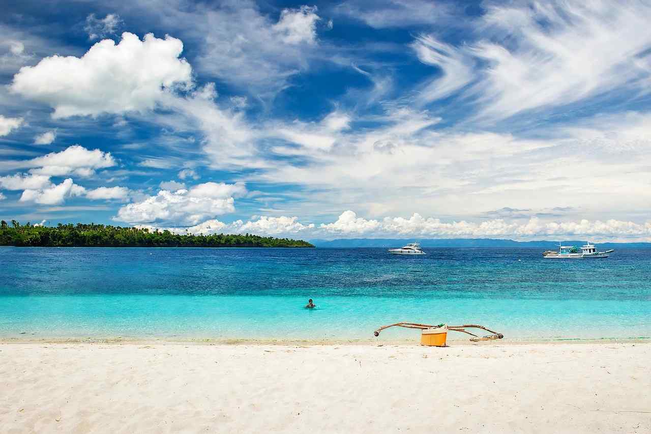 White sand and turquoise shallows on a Camotes Islands beach in Cebu Province, with an outrigger boat on the shore and boats anchored offshore