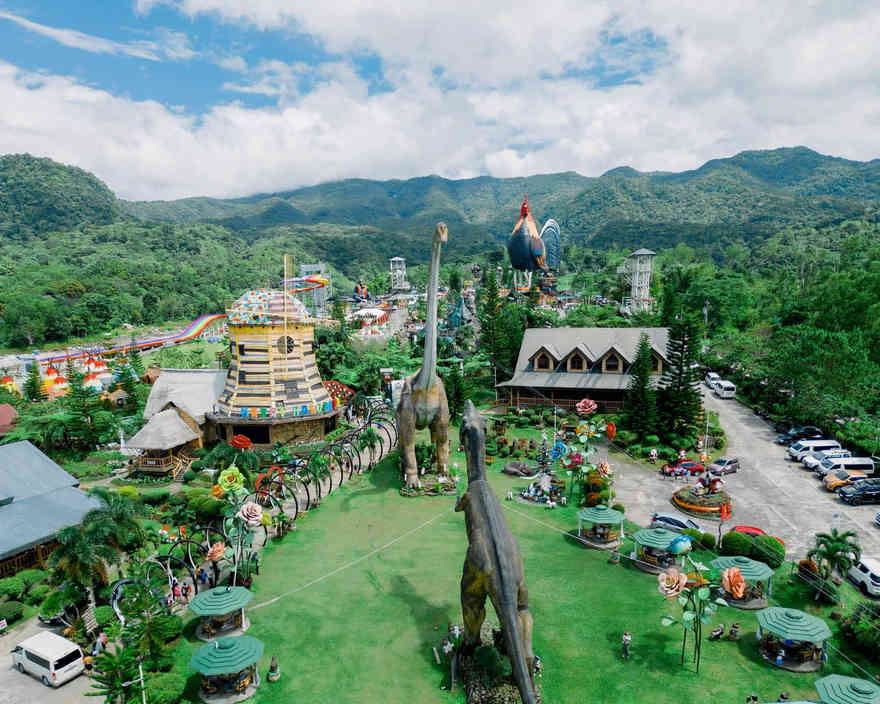 Aerial view of Campuestohan Highland Resort in Talisay, Negros Occidental, with dinosaur statues on a lawn, the giant rooster hotel, a rainbow water slide and forested hills behind