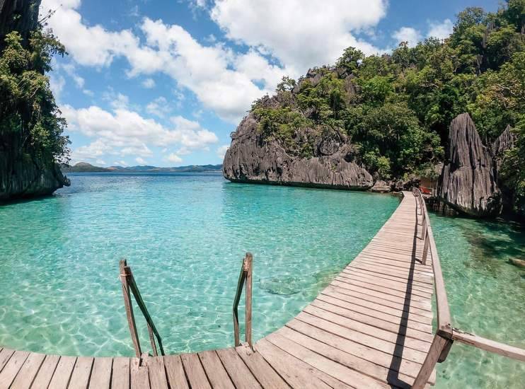 A wooden boardwalk with a swimming ladder over clear turquoise water, beside limestone cliffs on Coron Island, Palawan
