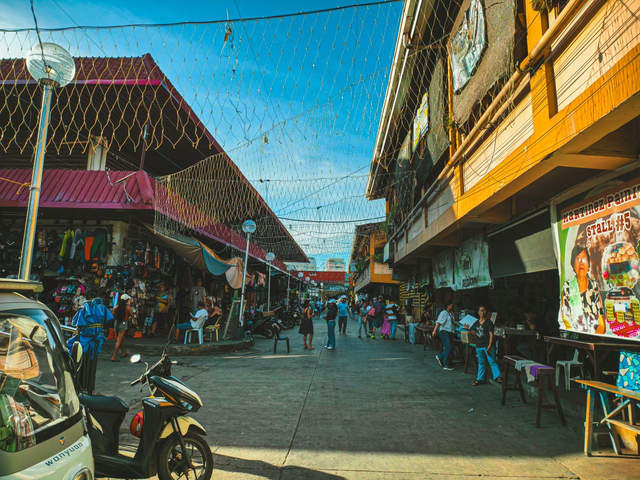 Busy market street lined with clothing stalls and small eateries under a canopy of netting on a sunny day