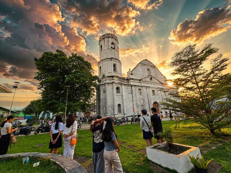 Visitors on a grassy lawn in front of a white stone church with a clock tower, under a sunset sky
