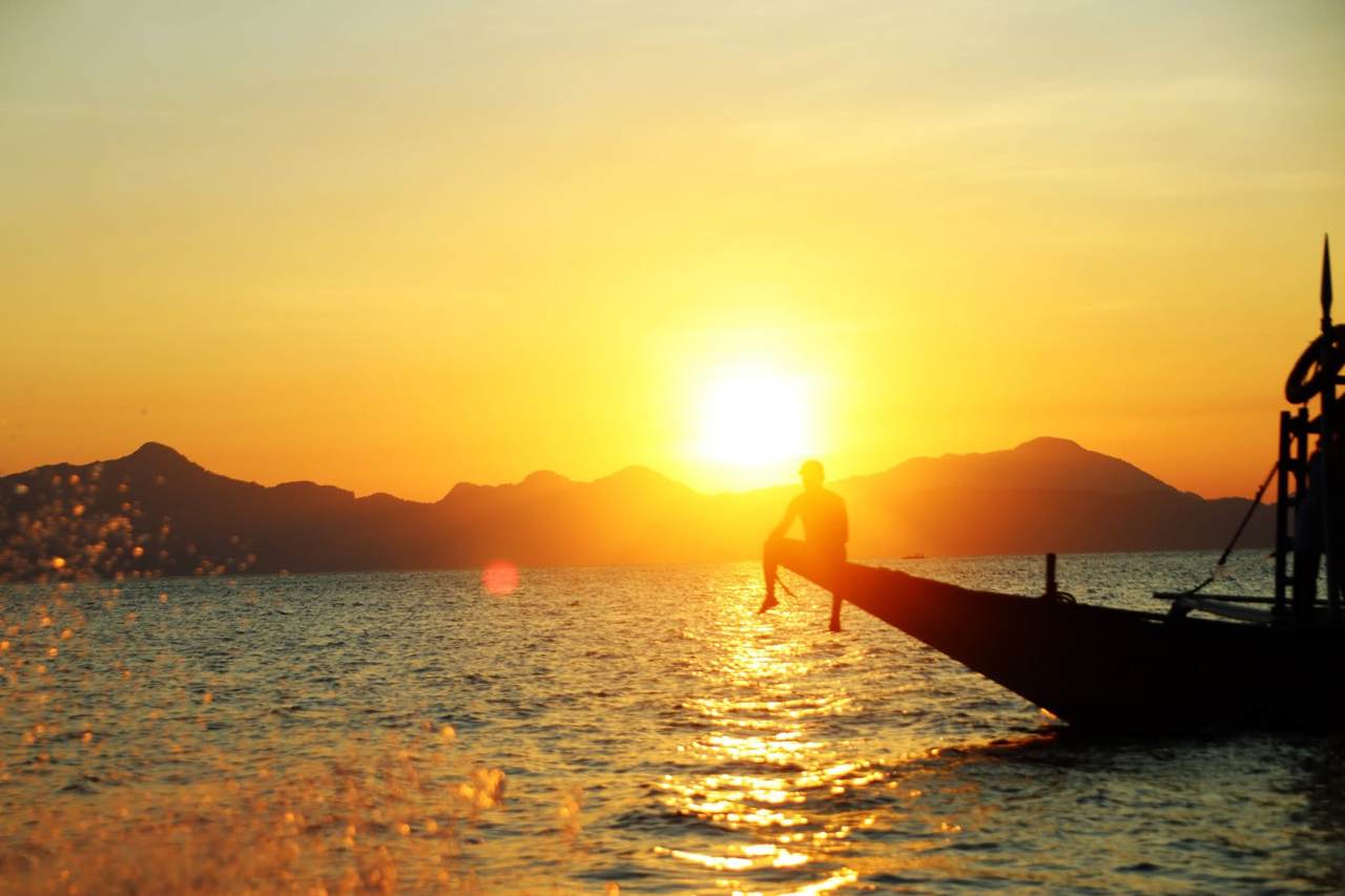A man sitting on the bow of an outrigger boat at sunset, with the sun setting behind the island hills of Bacuit Bay, El Nido