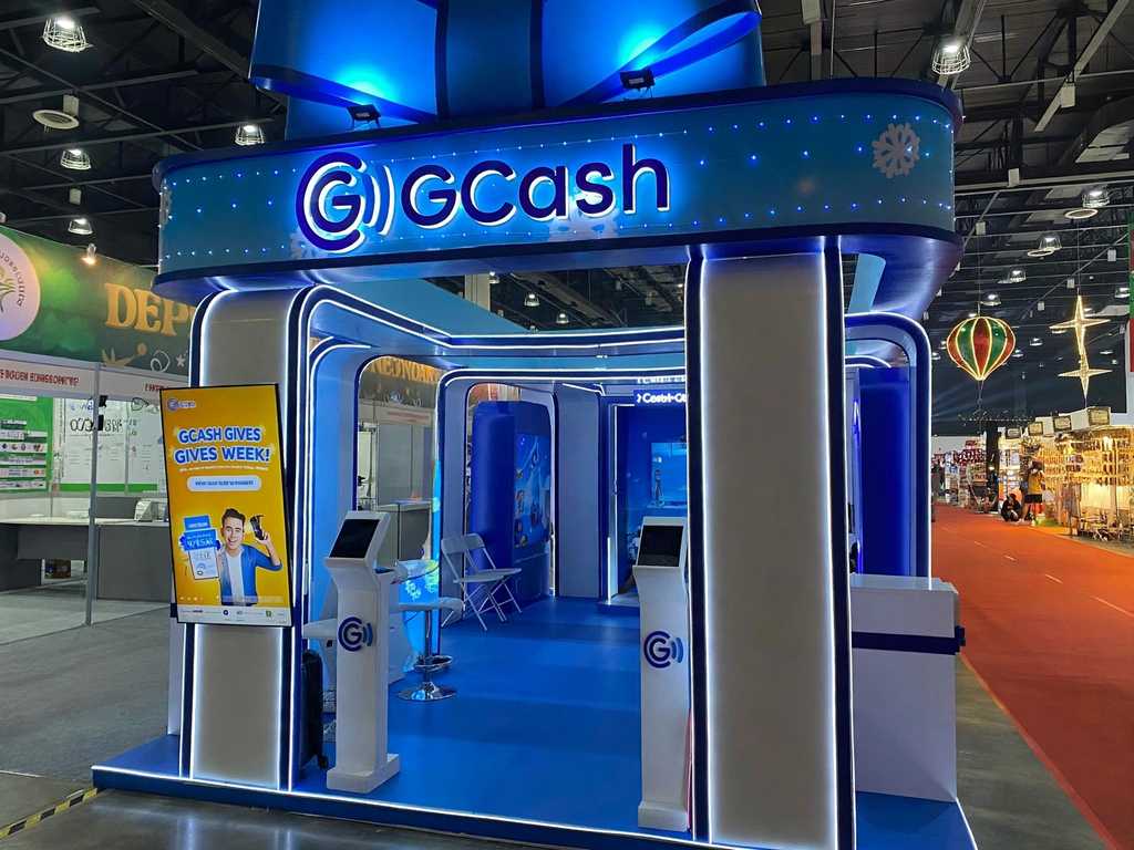 A blue-lit GCash booth at a trade fair, with self-service kiosks and a GCash Gives Week poster at the entrance