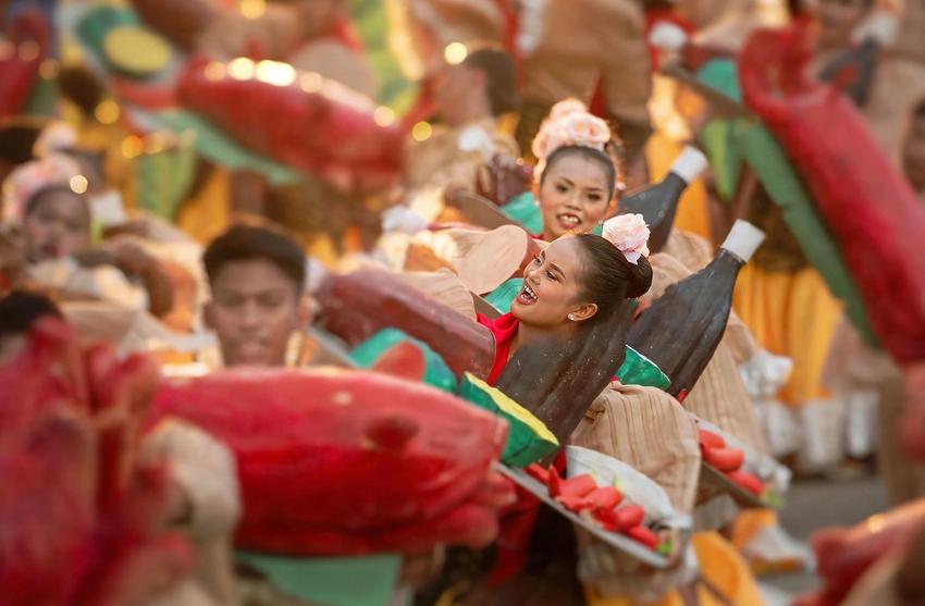 Festival dancers in red and brown costumes carry large red lechon props and bottle-shaped decorations along a street parade