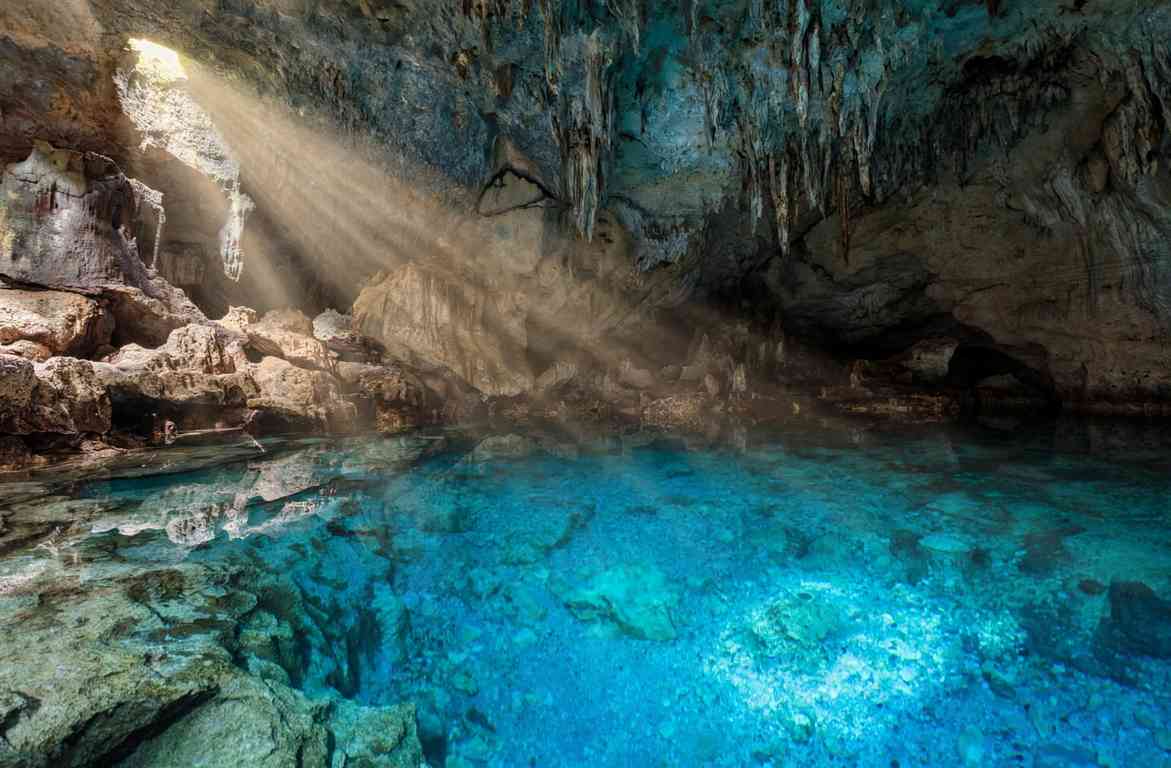 Sunbeams falling through an opening in the roof of Hinagdanan Cave onto a clear turquoise pool ringed by limestone and hanging stalactites