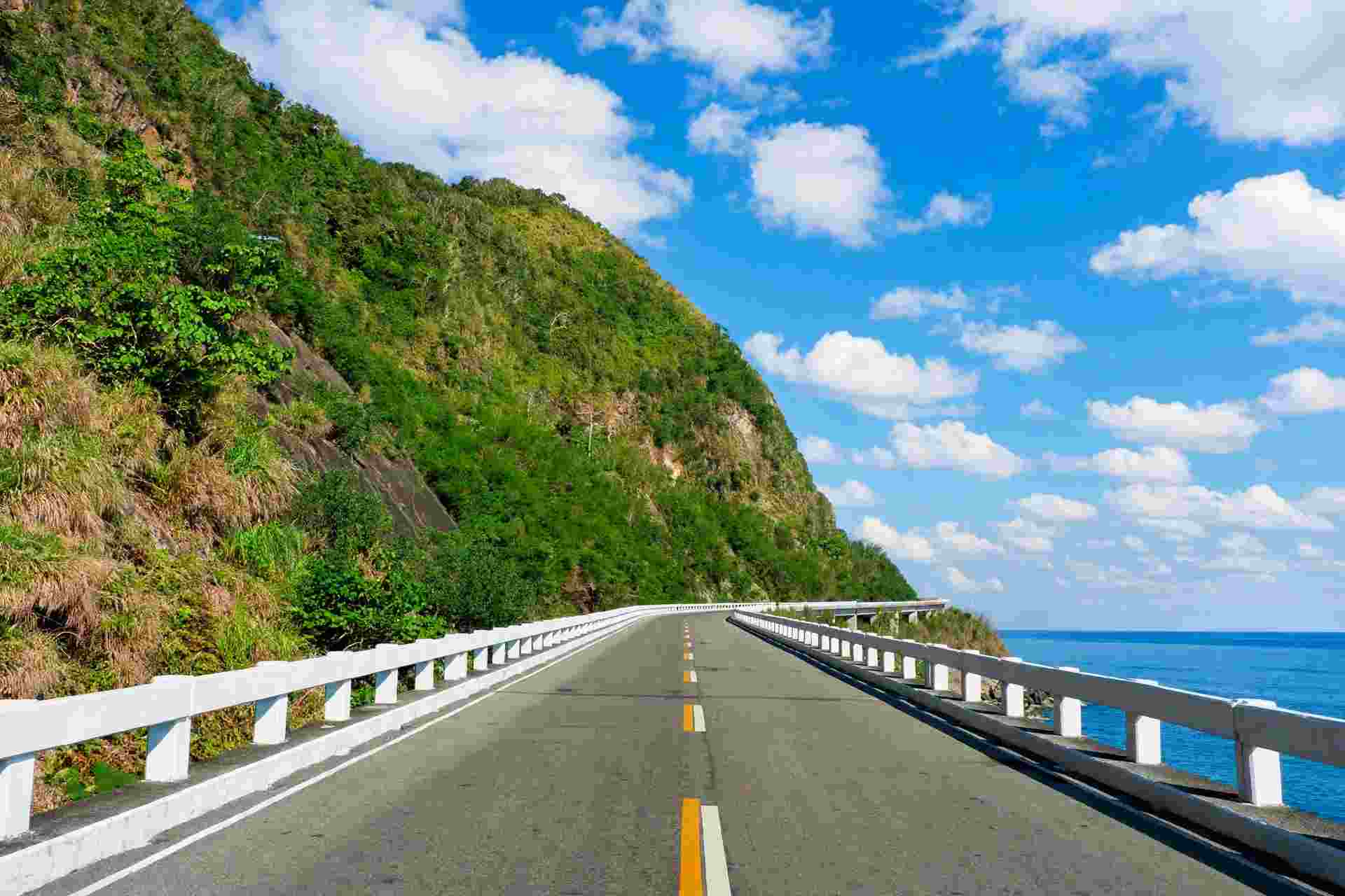 A two-lane coastal road with white guardrails curving along a green cliff above the sea under a bright blue sky