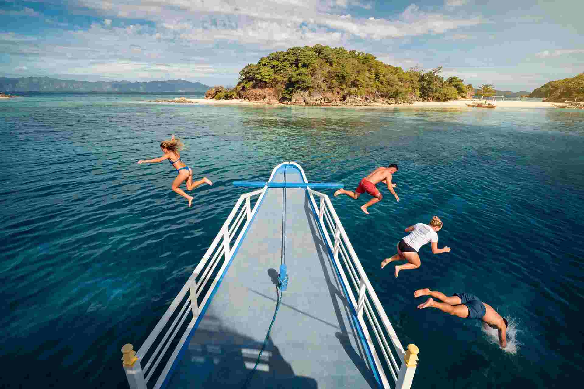 Four people jumping off the bow of a white-railed boat into clear blue water, with a forested island and a pale sandbar in the background