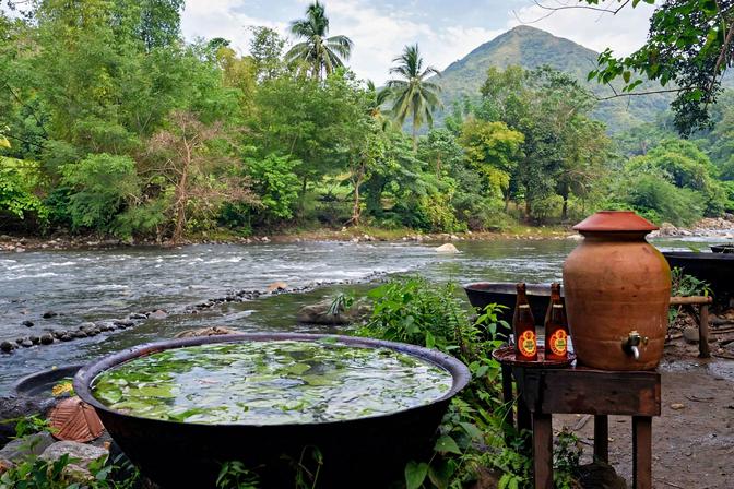 Large iron kawa filled with water and floating leaves beside a rocky river in Tibiao, Antique, with a clay water jug and a forested mountain behind