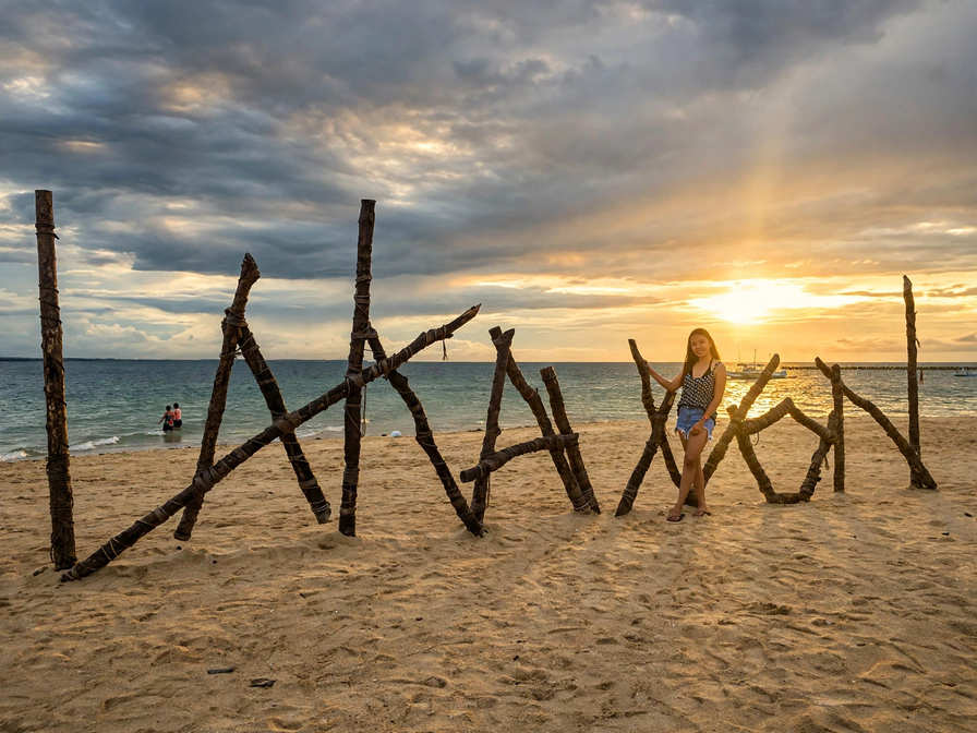 Driftwood letters spelling LAKAWON on the sand at sunset, with a visitor posing beside them and boats on the water behind
