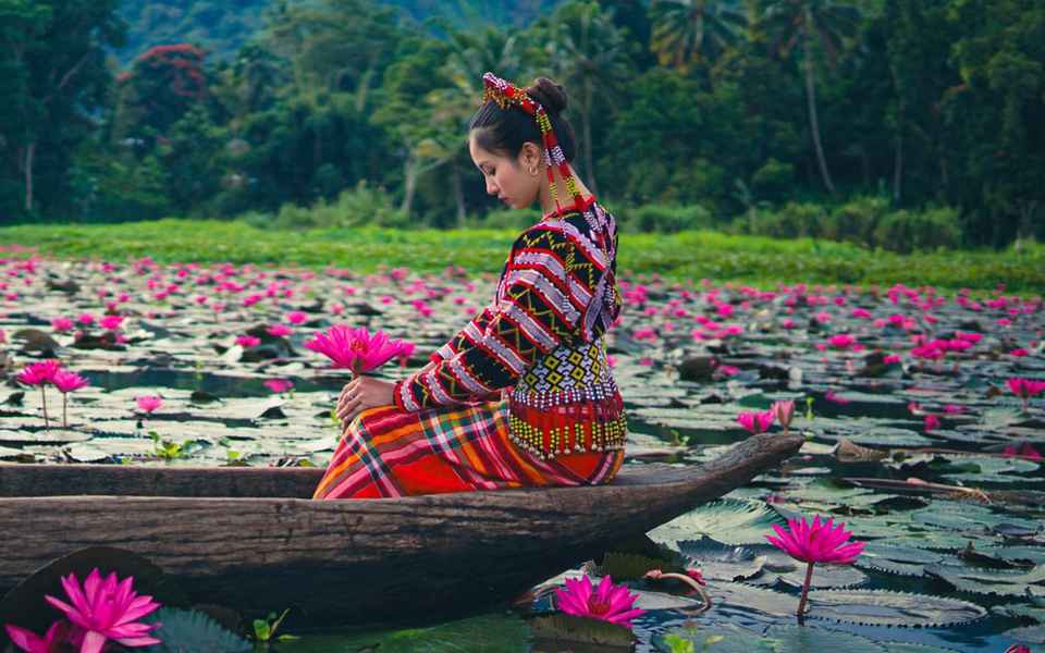 Woman in T'boli beaded headdress and woven attire seated on a wooden dugout canoe among pink lotus flowers on Lake Sebu