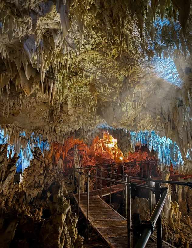 Wooden boardwalk with metal railings running through a cave, with stalactites hanging overhead lit in blue and orange, in Mambulod, La Libertad, Negros Oriental