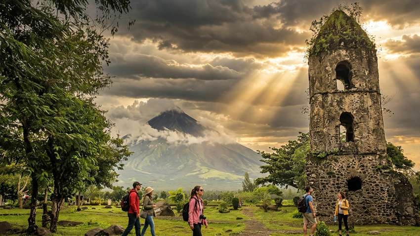 Hikers walking on the grass near the moss-covered Cagsawa Church bell tower with Mayon Volcano under clouds and sunbeams behind it