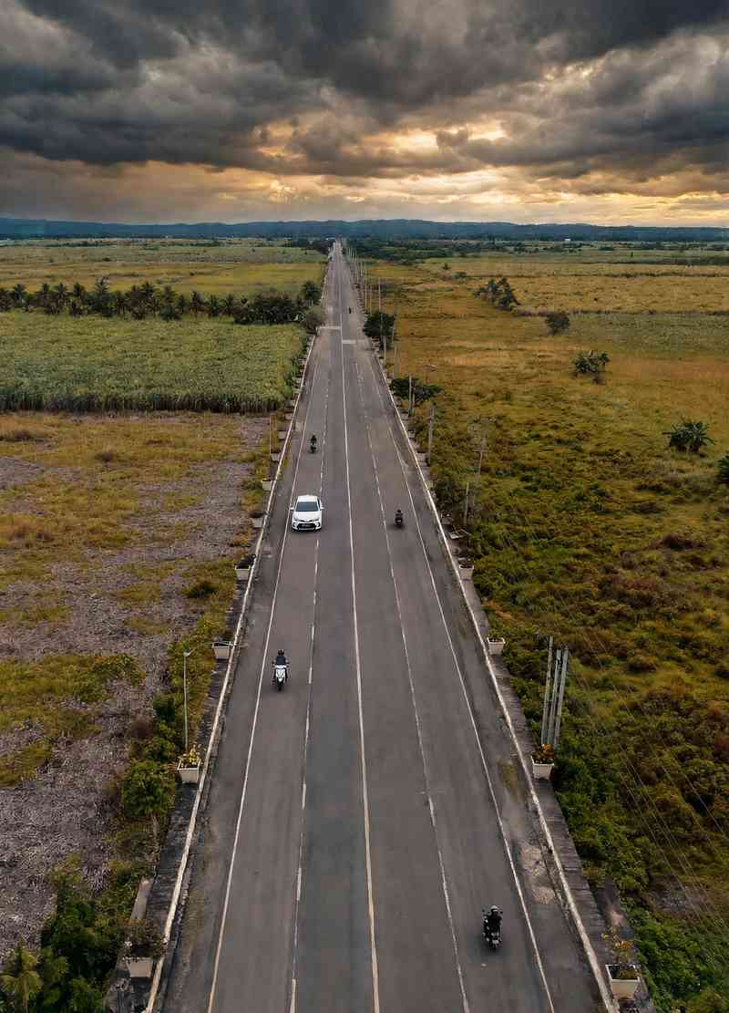 Aerial view of a long, straight road through flat sugarcane fields under heavy storm clouds, with motorbikes and a white car