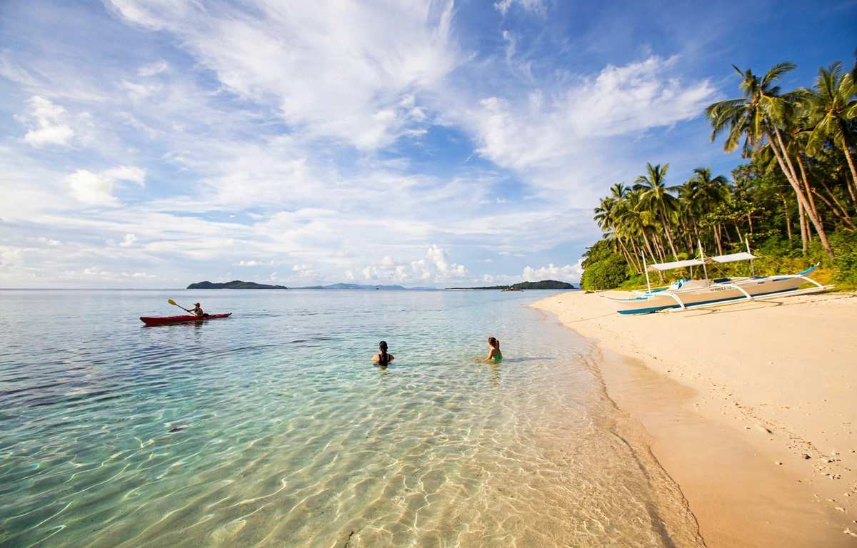 Two swimmers in clear shallow water near a white-sand beach in Palawan, with a kayaker offshore and an outrigger boat parked under coconut palms