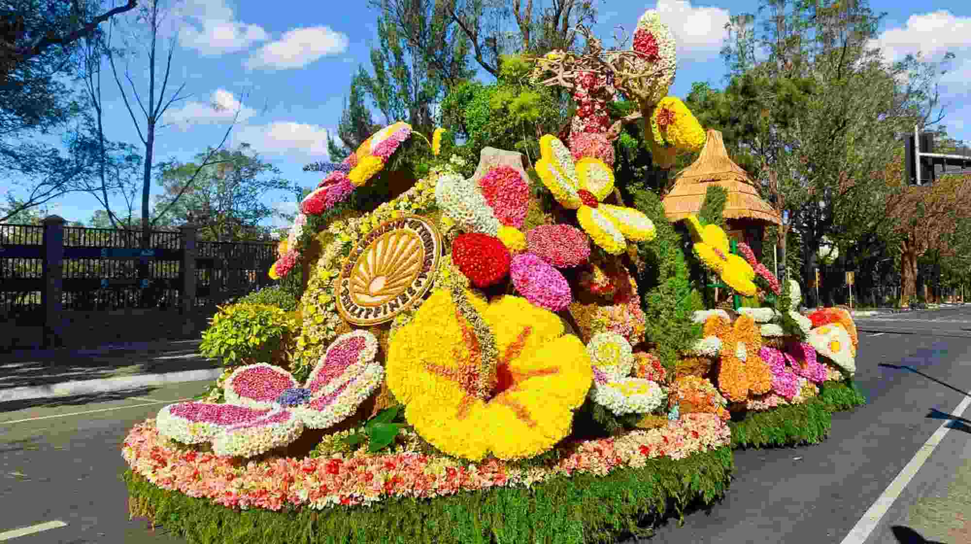 A float covered in yellow, red, pink and white chrysanthemums, with a golden sunburst emblem and a thatched nipa-style hut, on a tree-lined road