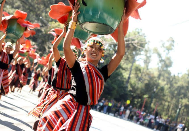 Panagbenga street dancer in a red and black striped Cordillera woven costume and beaded headdress, smiling as she holds a large flower-trimmed prop overhead during the parade in Baguio