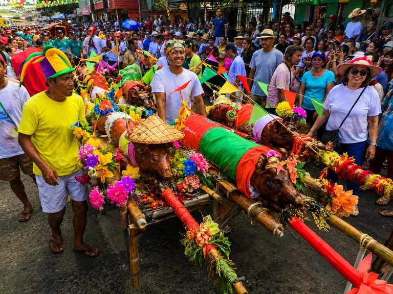 Whole roasted pigs dressed in a woven salakot, colourful cloth and paper flowers carried on bamboo litters through a packed street during Parada ng Lechon in Balayan, Batangas
