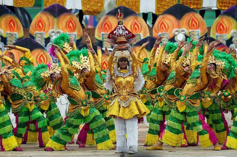 Pintados-Kasadyaan dancers in green and gold costumes with feathered headdresses surround a lead performer in gold raising an image of the Santo Niño