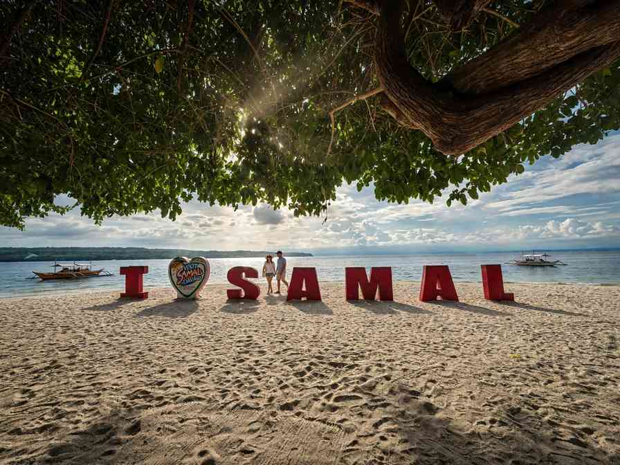 Red I Samal letters on a white-sand beach in Samal Island, shaded by a large tree, with bancas moored offshore