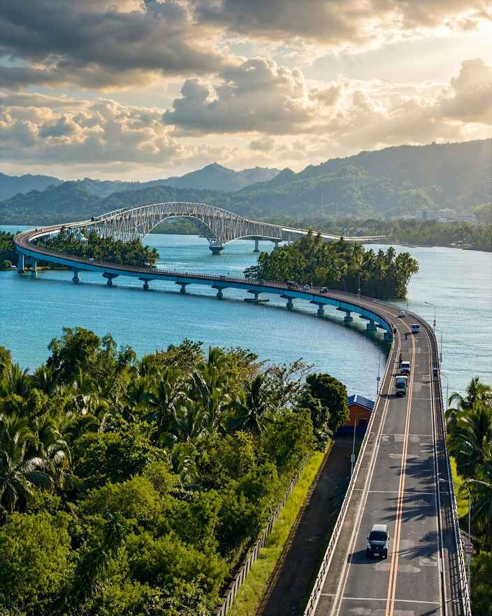 San Juanico Bridge curving across blue water between palm-covered shores in late afternoon light, with its arch truss in the distance and cars on the approach road