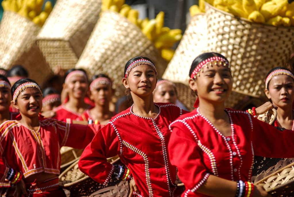 Young street dancers in red beaded costumes and headbands carrying woven baskets of yellow fruit during a festival parade