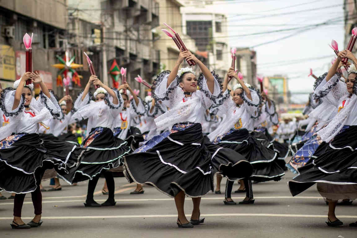 Female dancers in white lace blouses and swirling black skirts holding pink-tipped props overhead as they dance down a city street