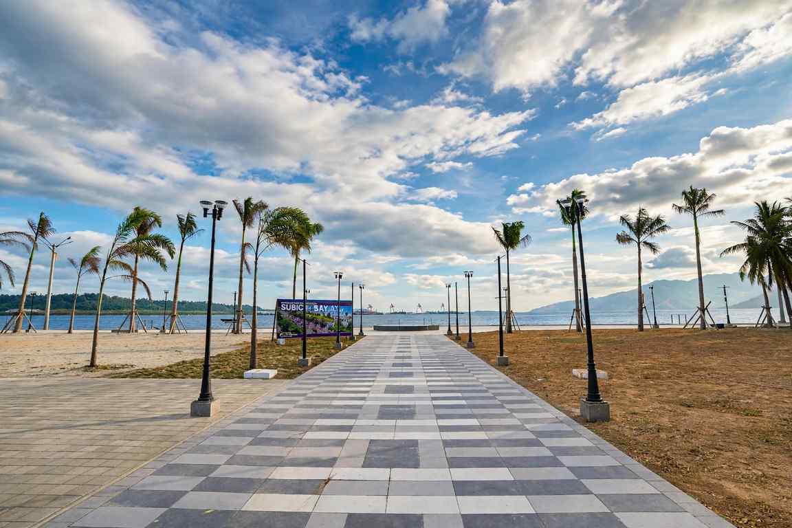 Paved waterfront promenade lined with palm trees and lamp posts leading to Subic Bay, with a Subic Bay sign, ships in the harbour and mountains under a cloudy blue sky