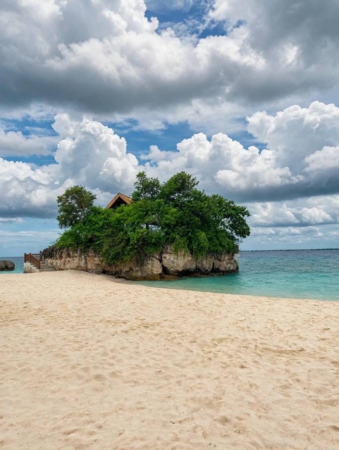 White sand beach with a tree-covered rock islet, a small hut on top and turquoise water under a cloudy sky