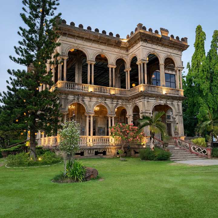 The Ruins in Talisay, Negros Occidental, at dusk: an arched Italianate mansion shell with a lit upper balcony, string lights in a pine tree and a green lawn