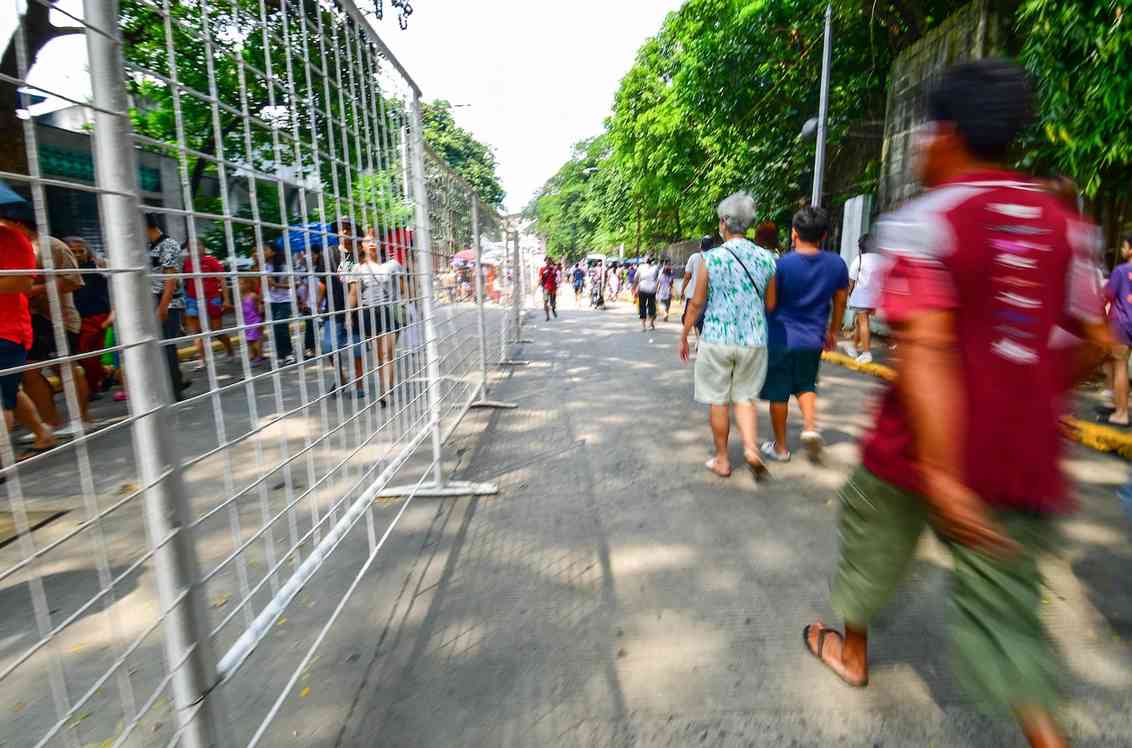 Families walking past metal crowd barriers on a tree-lined road outside a Manila cemetery during Undas